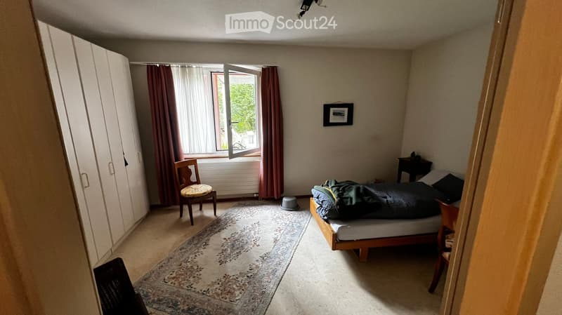 Bedroom with a bed, chair, wardrobe, small table, and a picture frame on the wall. The room has a window with curtains and a patterned rug on the floor.