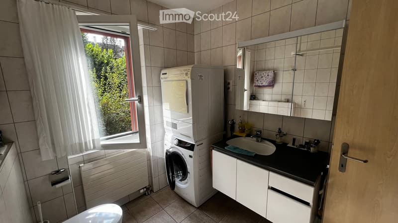 Bathroom with a sink, mirror, cabinet, washing machine, toilet, radiator, and a window with a curtain.