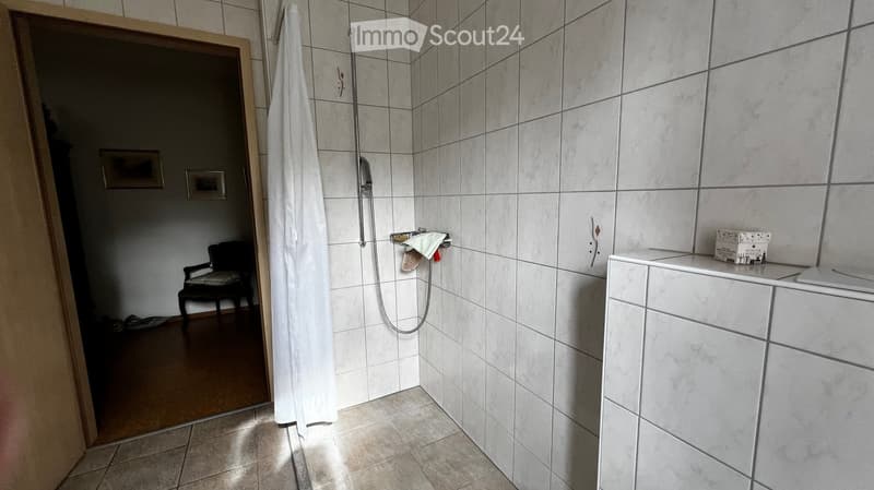 Modern bathroom with white tiled walls and floor, a shower with a curtain, a sink with a cabinet, a chair in the adjacent room, and a towel hanging on the wall.