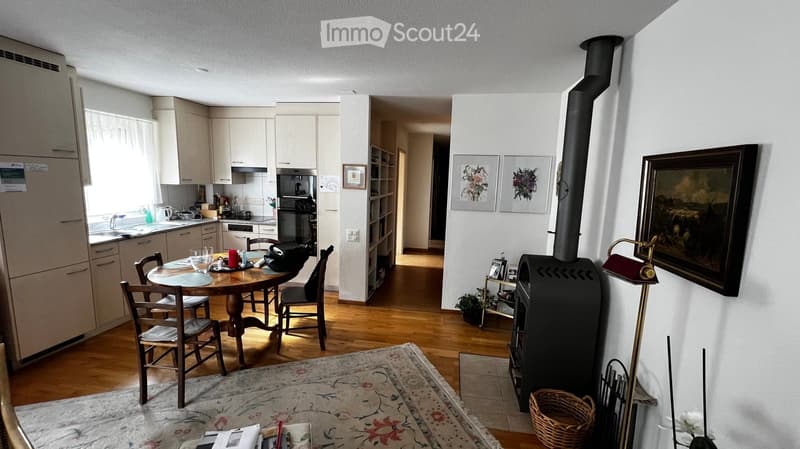 The image shows a well-lit, modern living and dining area with wooden flooring, a dining table with chairs, a kitchen area with cabinets and appliances, and a wood stove with a floor lamp nearby.