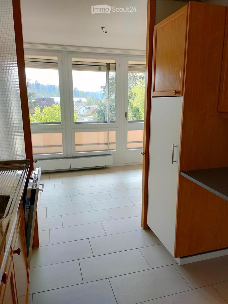 Empty kitchen with white tiled floor, wooden cabinets, and large windows offering a view of the outdoors.