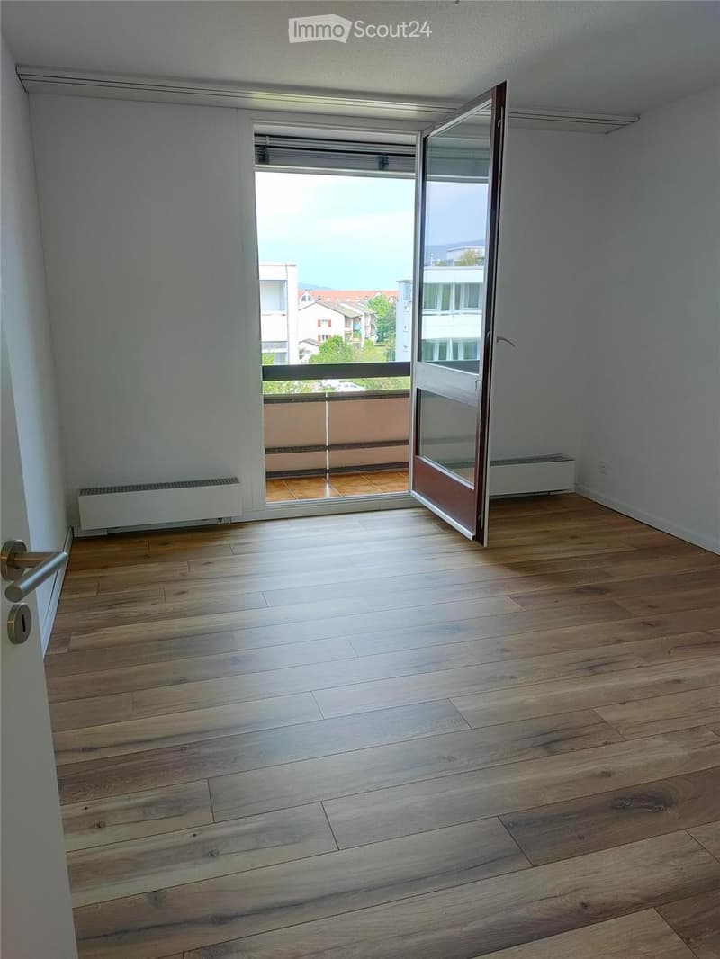 Empty room with wooden flooring, white walls, and an open sliding door leading to a balcony with a view of buildings and greenery.