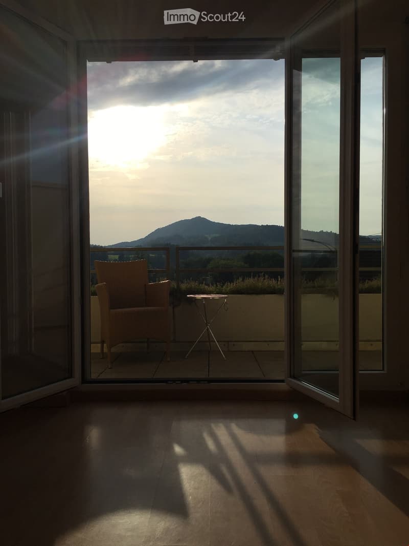 Balcony with a wicker chair and small table, overlooking a scenic mountain view during sunset.