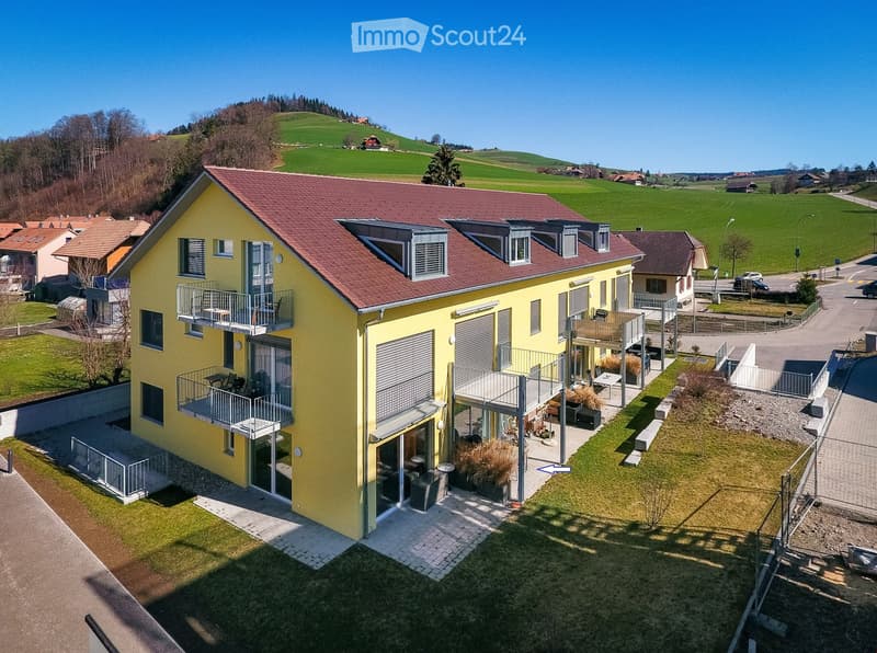 A modern yellow house with red-tiled roof, multiple balconies, and outdoor seating areas, surrounded by green fields and other houses.