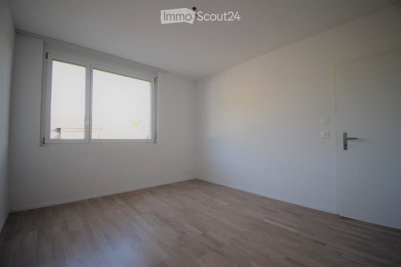 Empty room with light-colored walls, wooden flooring, and a large window with white frames. The room has a white door and visible electrical outlets.