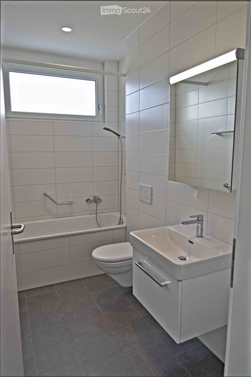 Modern bathroom with white tiles, a bathtub, toilet, sink, and a large mirror above the sink.