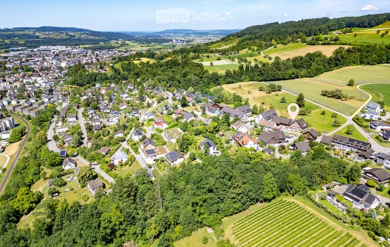Aerial view of a residential area with numerous houses, surrounded by green fields, forests, and distant hills.