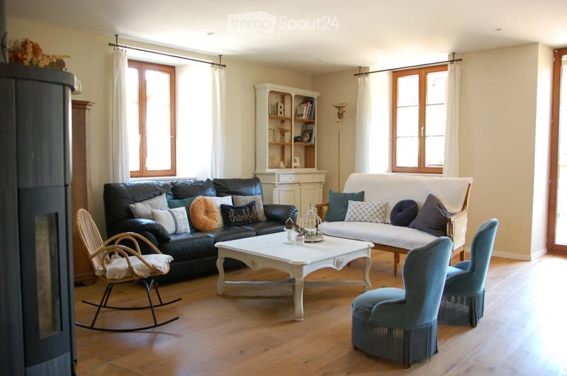 Bright living room with wooden flooring, large windows, a black leather sofa, a white coffee table, a rocking chair, and a blue armchair.
