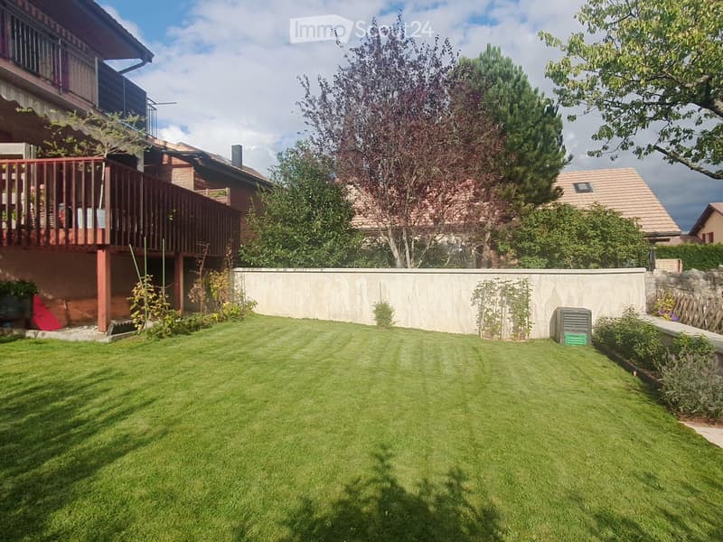 Well-maintained lawn with trimmed grass, surrounded by trees, shrubs, and a low concrete wall. A wooden fence and part of a house with a balcony are visible in the background.