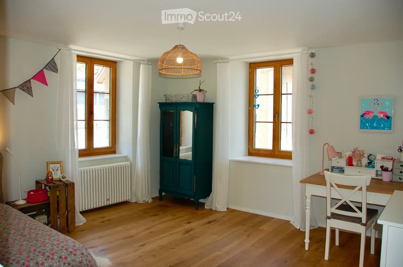 Cozy room with wooden flooring, large windows with white curtains, a bed with a floral duvet, a wooden chair and table, a dark green cabinet, a hanging pendant light, and decorative items including a picture frame and a plant on the windowsill.