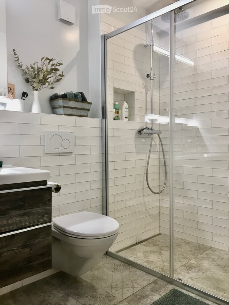 Modern bathroom with white tiles, a glass-enclosed shower, a toilet, a sink with a dark wooden cabinet, and decorative items on the windowsill.