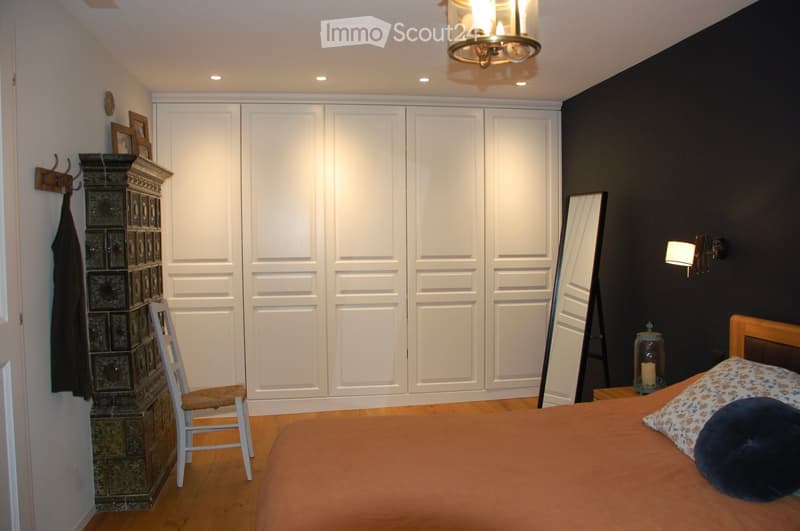 Bedroom with a large bed, a white wardrobe, a wooden chair, a mirror, and a candle holder on a bedside table.