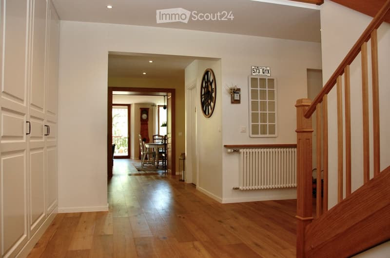 Spacious hallway with wooden flooring, white walls, and a staircase leading upwards. Features a radiator, a wall-mounted clock, and a framed picture. A dining area is visible in the background with a table and chairs.