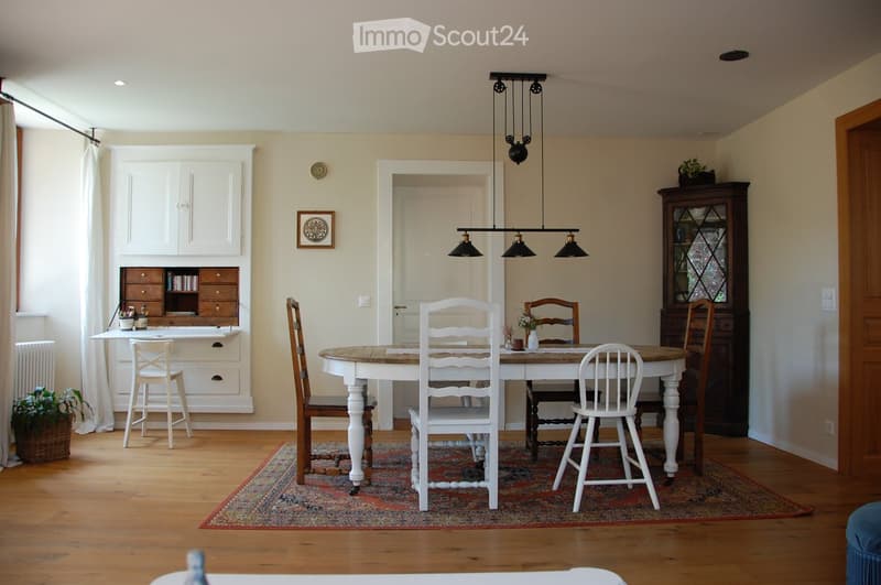 Bright dining area with wooden floor, large wooden dining table surrounded by white and wooden chairs, a hanging light fixture, a white cabinet with drawers, a small desk with a chair, and a dark wooden cabinet with glass doors.