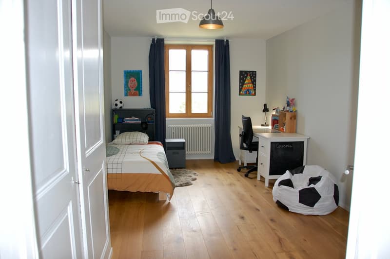 Well-lit bedroom with wooden flooring, a bed with orange and white bedding, a desk with a chair, a bookshelf, a beanie bag, and a window with dark curtains.