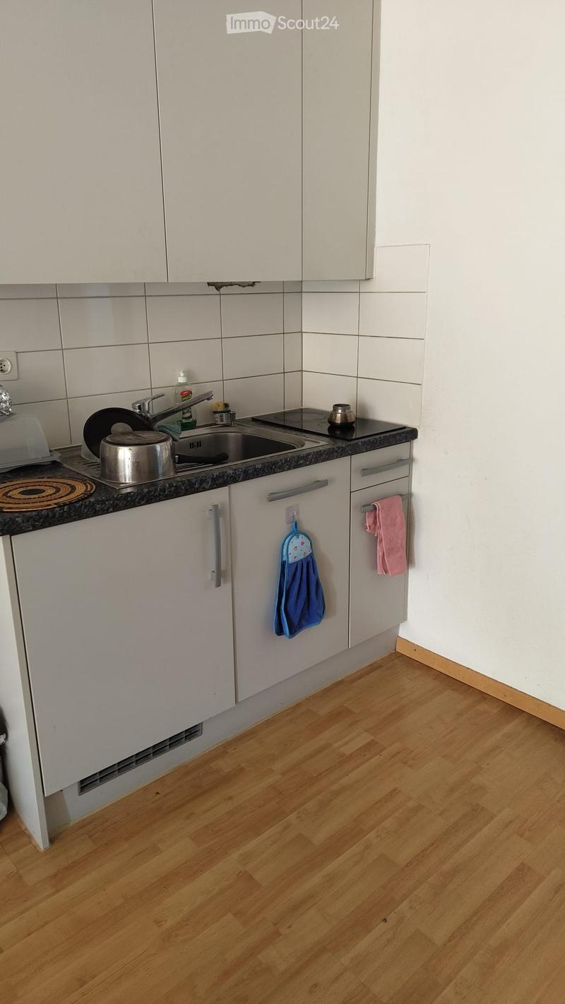 Modern kitchen with light-colored cabinets, wooden flooring, and a black countertop. Visible items include a sink, faucet, oven, pot, and towels.