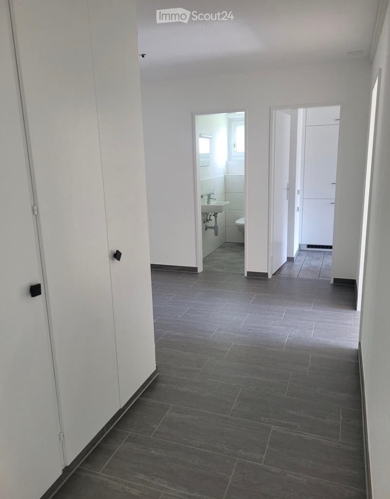Empty hallway with grey tiled floor, white walls, and two open doors leading to a bathroom and another room.