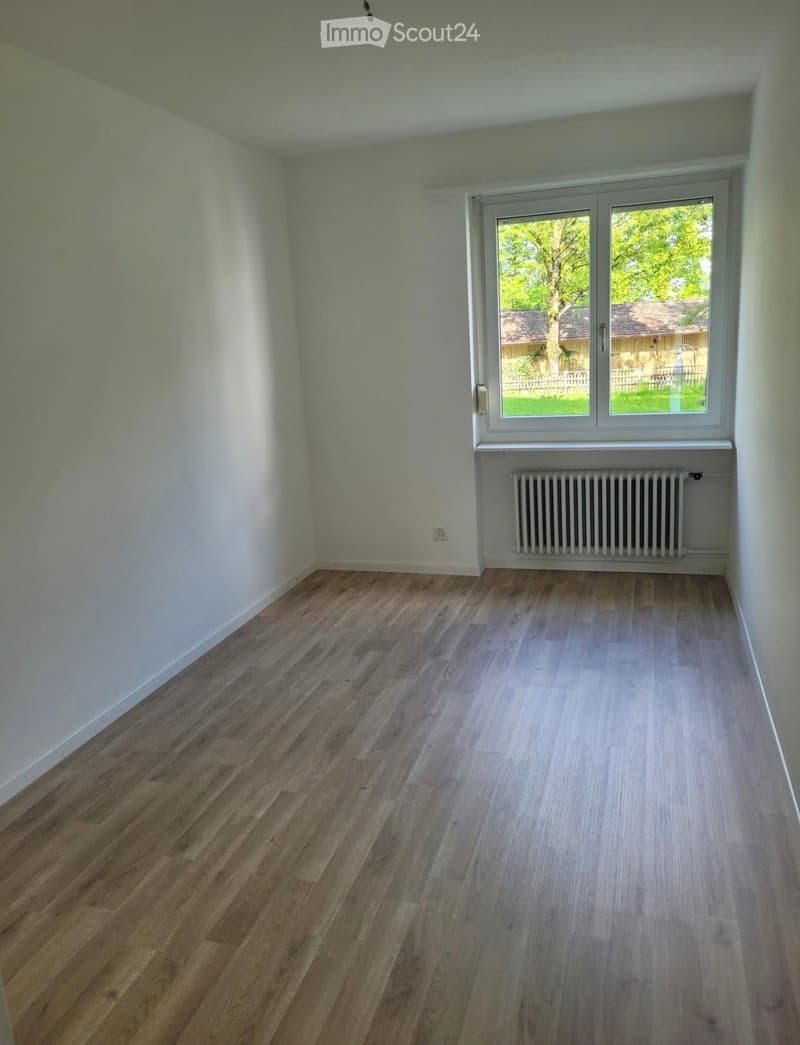 Empty room with wooden flooring, white walls, a window with a radiator below, and a view of a garden outside.