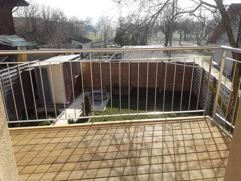 Balcony with wooden decking and metal railing, overlooking a fenced yard with a small pool and surrounding structures.