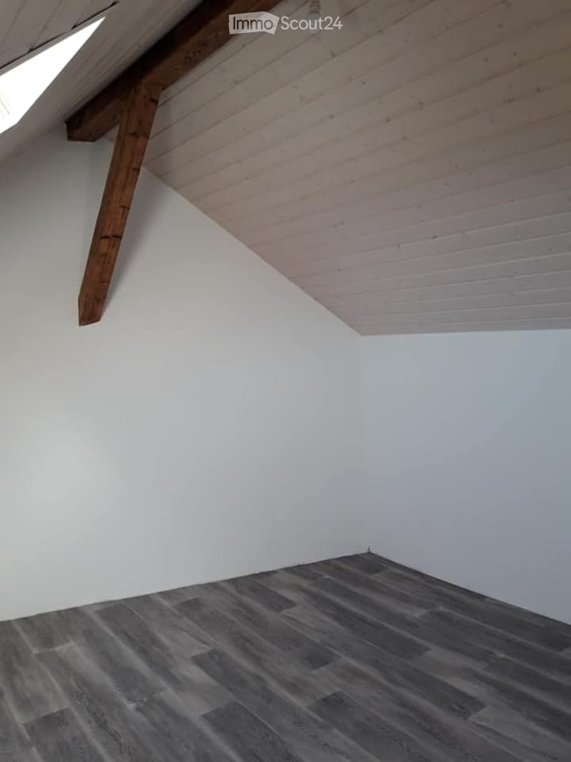 Empty attic room with wooden slanted ceiling, exposed wooden beam, and dark grey wooden flooring.