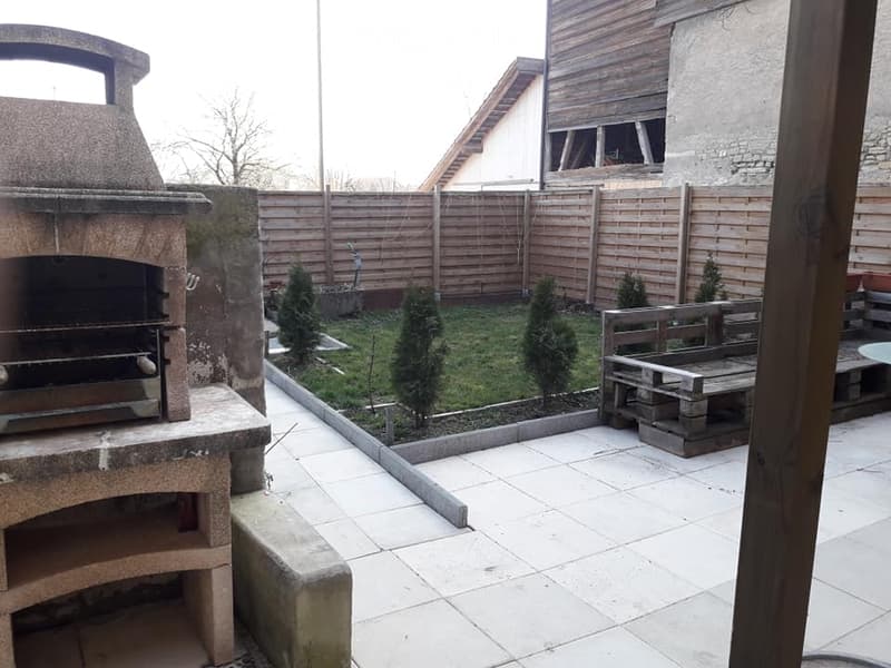 A small, enclosed garden with a tiled patio, a wooden fence, a stone pizza oven, a small seating area, and several small evergreen trees.