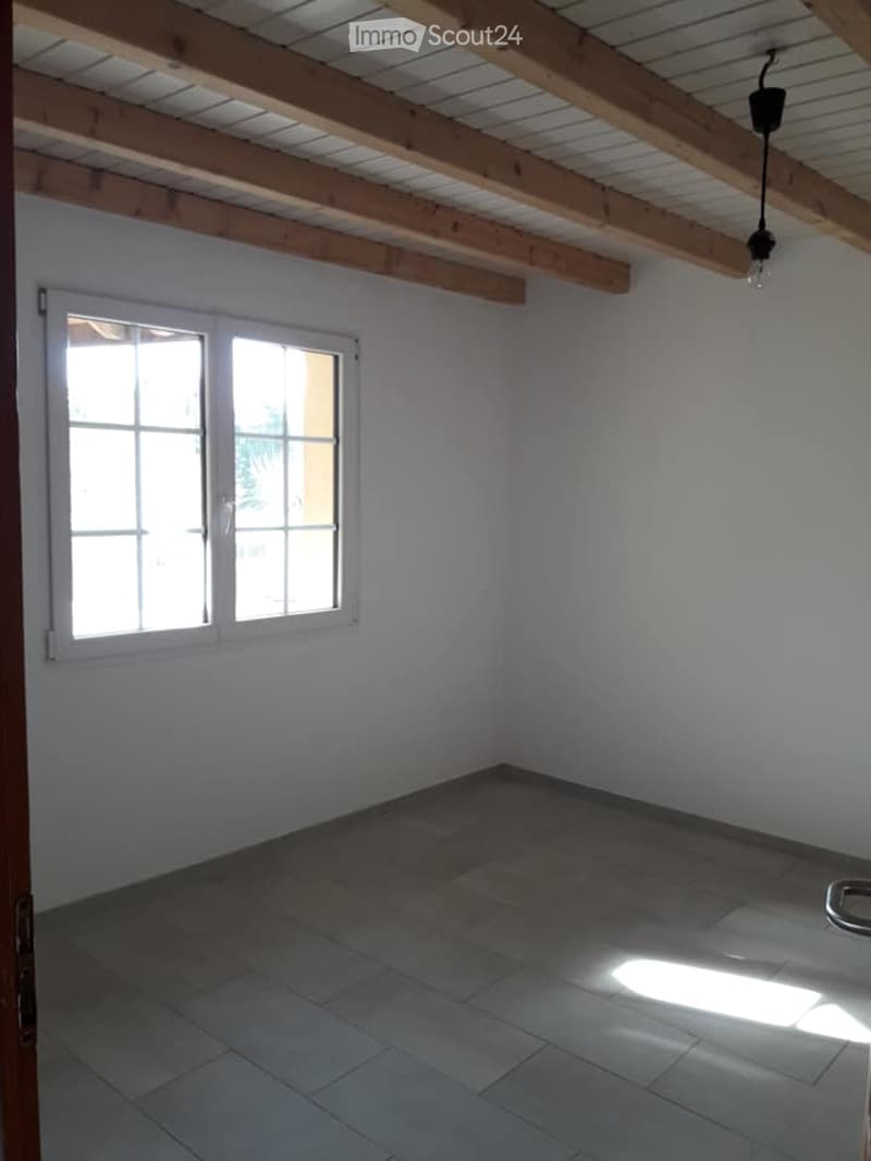 Empty room with exposed wooden beams on the ceiling, white walls, tiled floor, and a window letting in natural light.