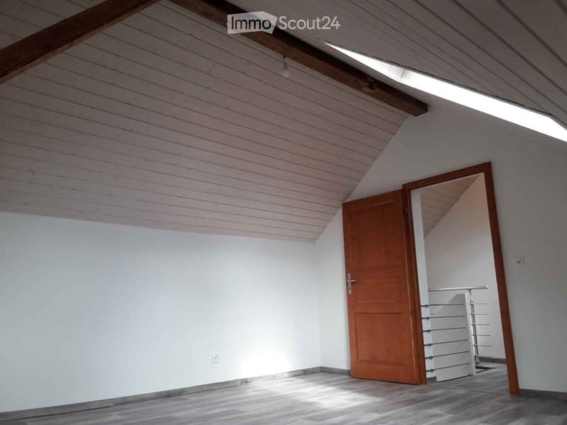 Empty attic room with wooden ceiling beams, white paneled walls, and a wooden door leading to a staircase. The floor is light-colored wood, and natural light streams in from a skylight.