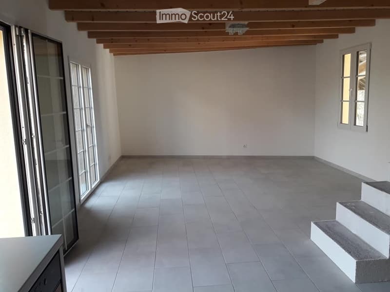 Empty room with tiled floor, white walls, exposed wooden ceiling beams, large glass sliding doors, and a small staircase leading upwards.