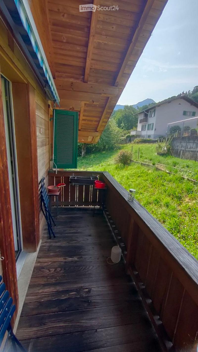 Wooden balcony with a view of green fields and distant mountains, featuring a small table and chairs, and a barbecue.