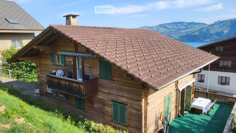 A two-story wooden house with a brown tiled roof, green shutters, and a balcony overlooking a scenic lake and mountain view.