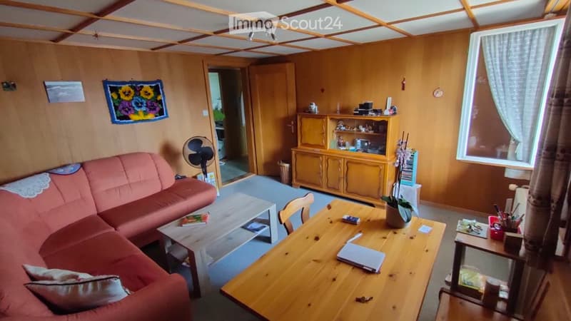 A cozy living room with wooden walls, a red sofa, a coffee table, a wooden dining table, a fan, a cabinet, and a plant. The room has a warm, inviting atmosphere with natural light coming through a window.