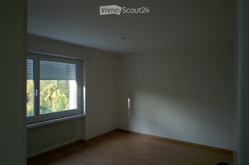 Empty room with wooden flooring, white walls, and a large window with blinds, allowing natural light to enter.