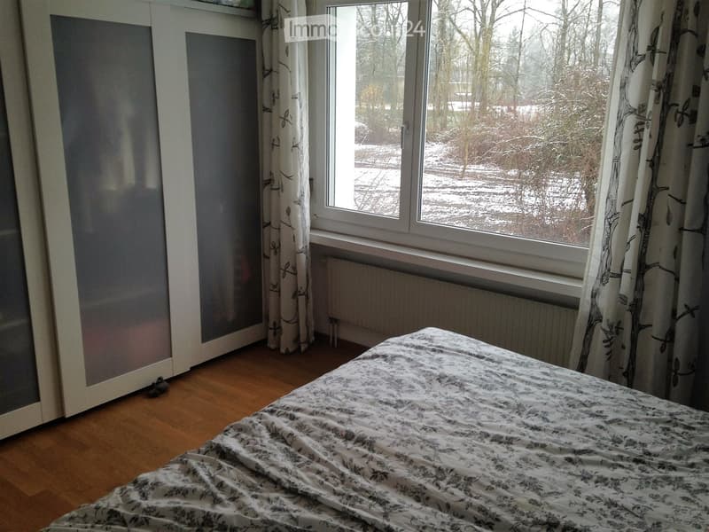 Bedroom with wooden floor, large window with curtains, bed with floral bedding, and a wardrobe with mirrored doors.