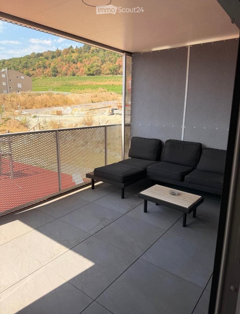 Modern balcony with grey tiled floor, black sectional sofa, small wooden table, and scenic countryside view.