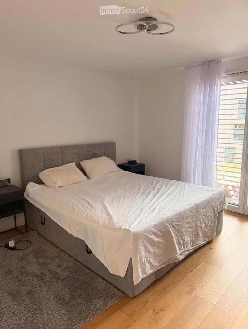 Modern bedroom with a large bed, white bedding, nightstands, and a ceiling light fixture. The room features wooden flooring and a large window with curtains.