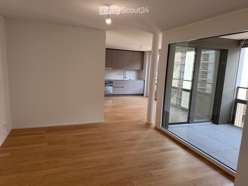 Empty living space with wooden flooring, white walls, and a sliding glass door leading to a balcony. The kitchen area is visible in the background with cabinets and a sink.