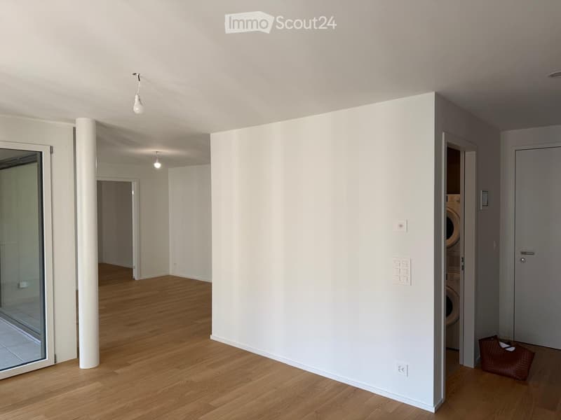 Empty room with wooden flooring, white walls, and a visible laundry area in the background.