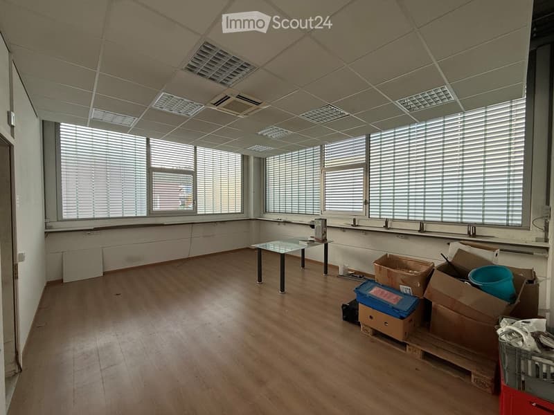 A spacious, unfurnished room with wooden flooring, large windows with blinds, and a glass table with a few items on it. Cardboard boxes and a blue container are stacked in the corner.
