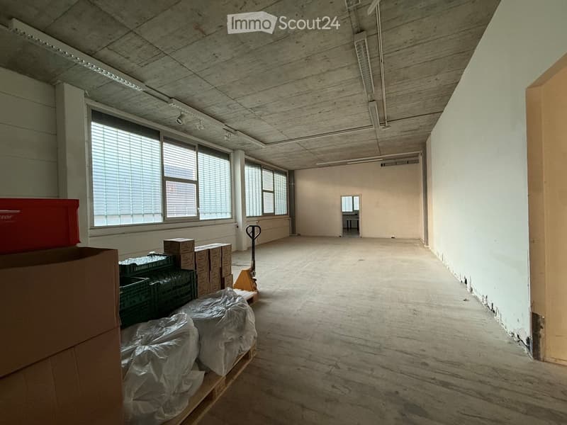 Empty room with bare walls, concrete floor, large windows, and some stacked boxes and crates in the corner.