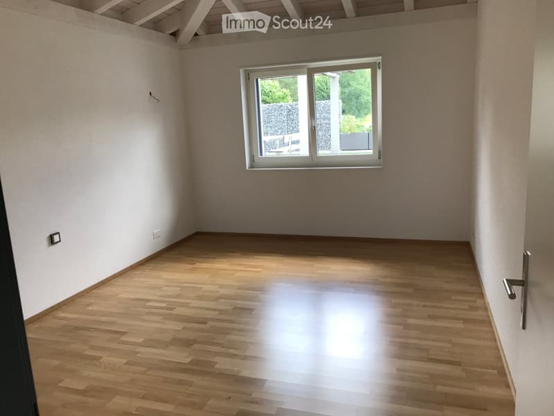 Empty room with light wooden flooring and white walls, featuring a large window with a view of greenery outside.