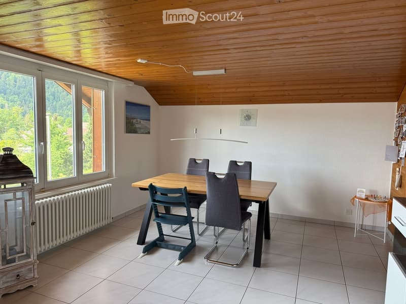 Dining area with a wooden table, four chairs, a radiator, a lantern, and a clock on the wall. Large windows offer a view of greenery.