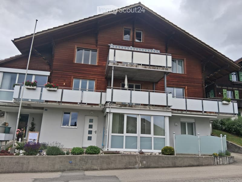 Two-story wooden house with multiple balconies, large windows, and flower boxes. The lower level has a white facade with a central entrance and large glass doors.
