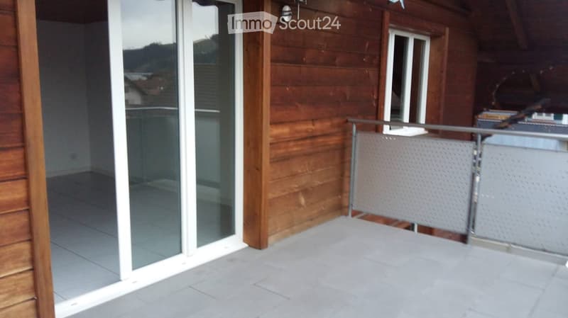 Balcony with grey tiled floor, glass sliding door, wooden railing, and a view of the surrounding area.