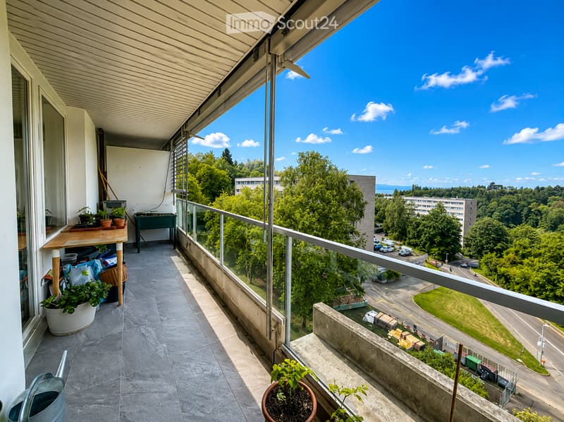 Balcony with tiled floor, glass railing, and potted plants. Offers a view of trees, buildings, and a road below.