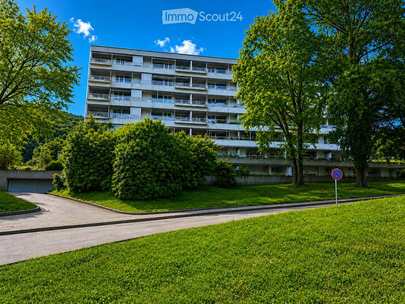 Multi-story residential building with balconies, surrounded by greenery and a paved pathway.