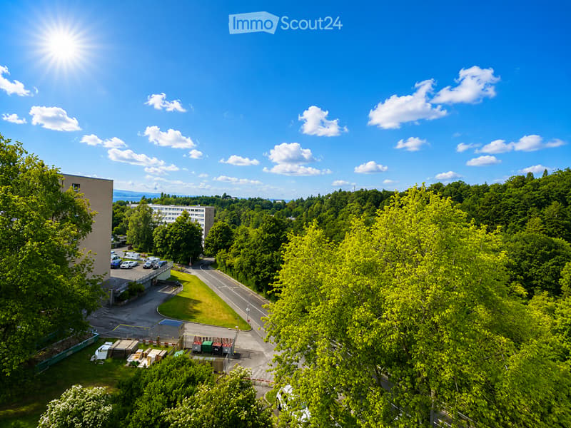 Aerial view of a modern building surrounded by lush green trees and a parking area with several cars. The scene includes a clear blue sky with scattered clouds and a bright sun shining from the left side.