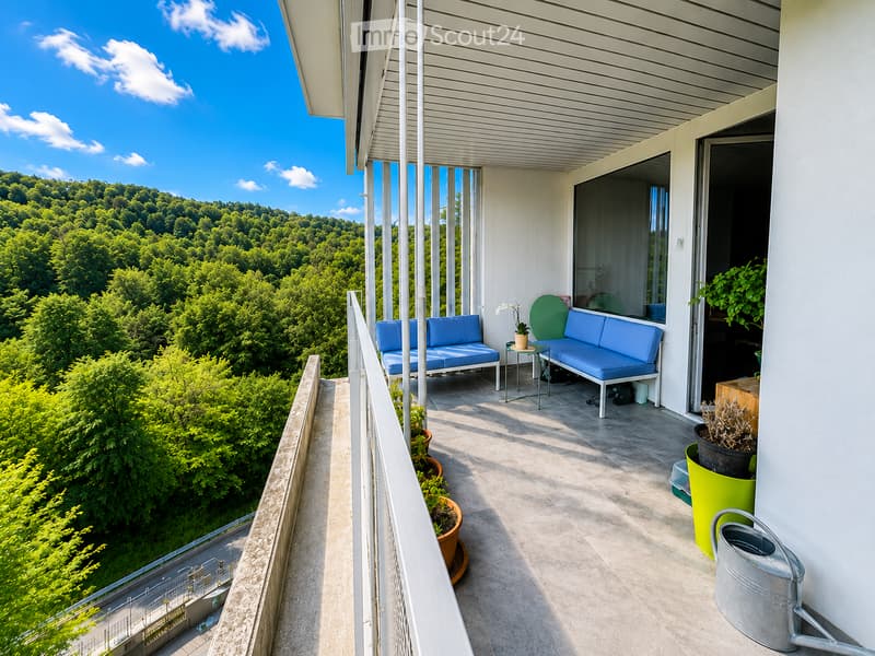 Balcony with modern furniture, including a blue sofa, small table, and chairs, overlooking a lush green landscape.