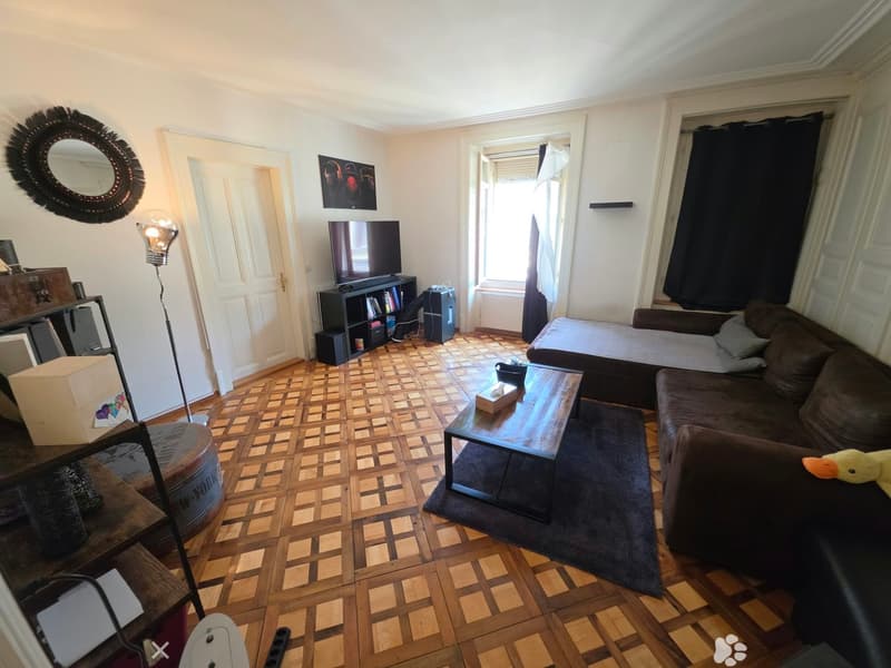 Living room with wooden parquet flooring, a dark brown sofa, a coffee table, a TV on a stand, a floor lamp, a round mirror, and a shelf with books and items.