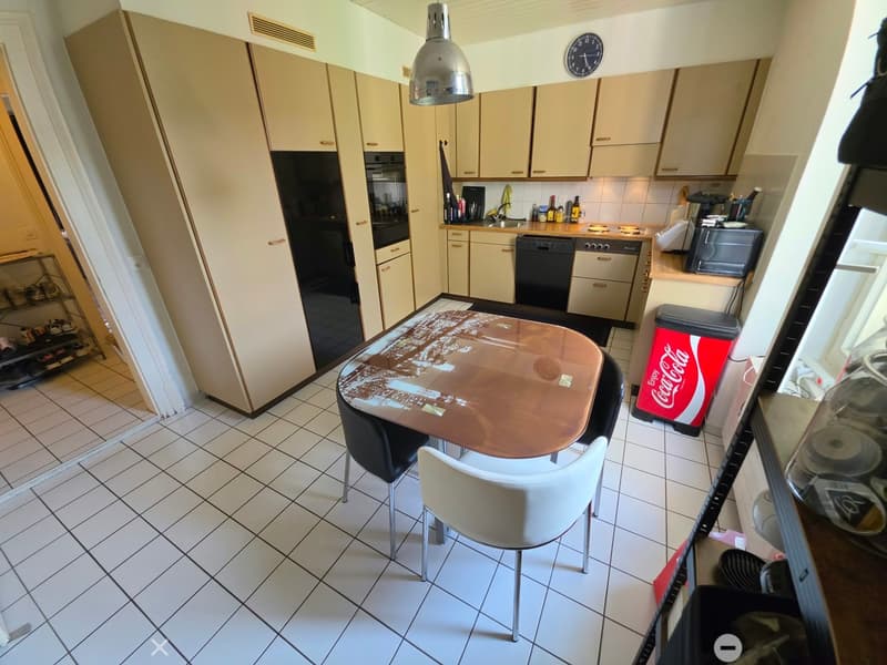 A modern kitchen with white cabinets, a central dining table with chairs, and various appliances including a refrigerator, oven, dishwasher, and microwave. The floor is tiled, and there is a Coca-Cola trash bin visible.