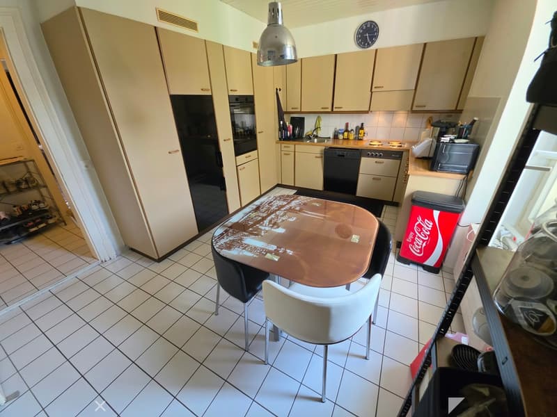 A modern kitchen with white cabinets, a central dining table with chairs, and various appliances including a refrigerator, oven, dishwasher, and microwave. The floor is tiled, and there is a Coca-Cola trash bin visible.